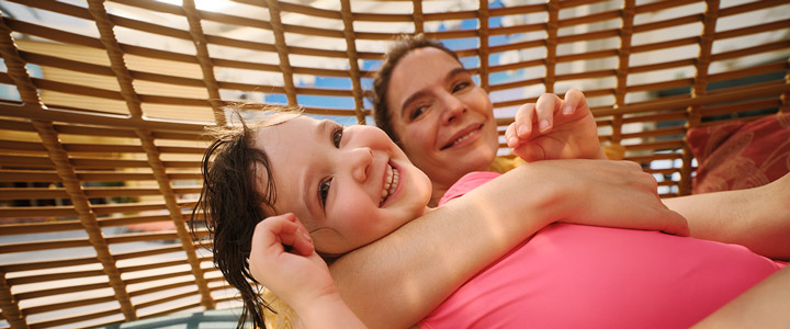 Fröhliches Mädchen mit pinkem Badeanzug lässt in der Umarmung mit ihrer Mutter die Seele am Pool baumeln.