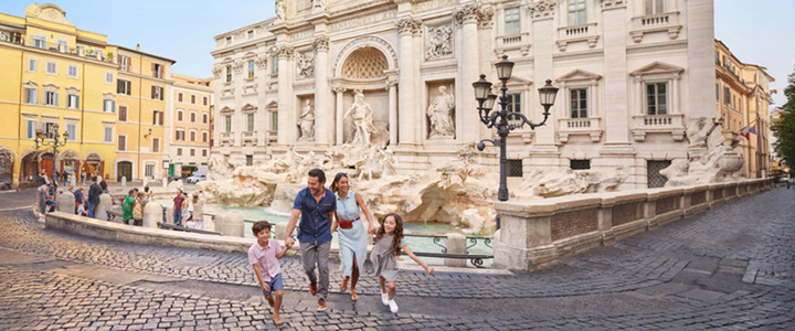 Ausgelassene Familie am Trevi-Brunnen in Rom im Vordergrund, dahinter fällt das Wasser über helle Marmorstatuen in warmes Licht.